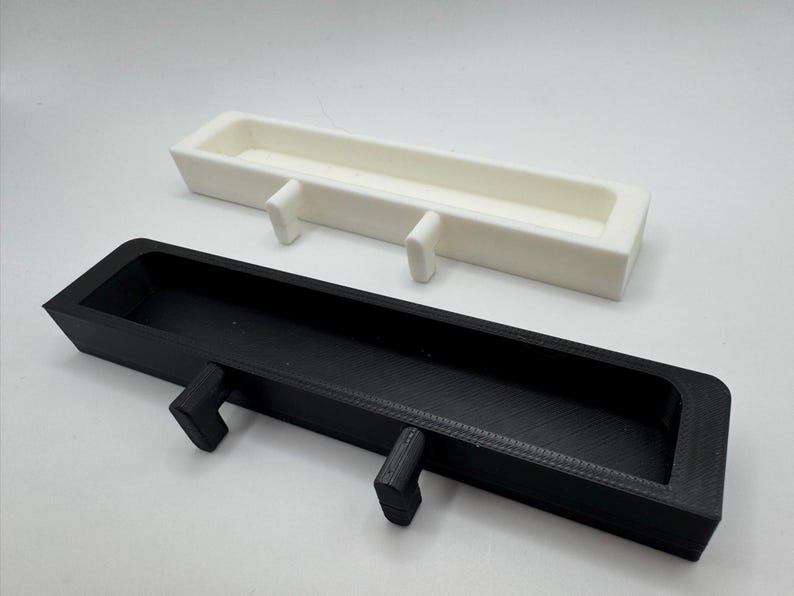May include: Two rectangular, open-top trays with integrated hooks. One tray is black, the other is white. Both have a similar design, with a long, narrow shape and two protruding hooks on the underside for hanging.
