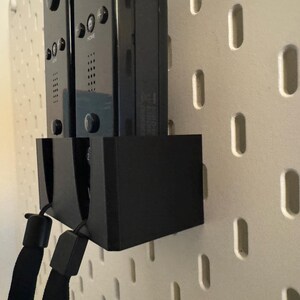 Puede incluir: Soporte negro para mandos de juego con dos mandos adjuntos, montado en una pared perforada blanca. El soporte tiene un dise&ntilde;o rectangular negro s&oacute;lido. Los mandos son negros con botones y correas.