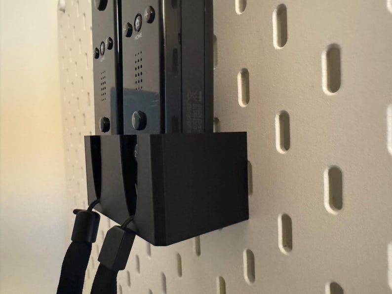 Puede incluir: Soporte negro para mandos de juego con dos mandos adjuntos, montado en una pared perforada blanca. El soporte tiene un dise&ntilde;o rectangular negro s&oacute;lido. Los mandos son negros con botones y correas.