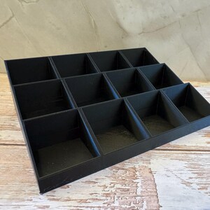 Peut inclure: Un organisateur noir &agrave; plusieurs niveaux avec douze compartiments. L'organisateur est fait d'un mat&eacute;riau noir mat et est con&ccedil;u pour contenir de petits objets. Les compartiments sont dispos&eacute;s en trois rang&eacute;es, chacune ayant une profondeur diff&eacute;rente.