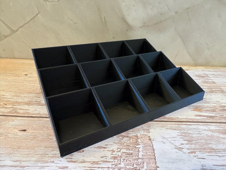 Peut inclure: Un organisateur noir &agrave; plusieurs niveaux avec douze compartiments. L'organisateur est fait d'un mat&eacute;riau noir mat et est con&ccedil;u pour contenir de petits objets. Les compartiments sont dispos&eacute;s en trois rang&eacute;es, chacune ayant une profondeur diff&eacute;rente.