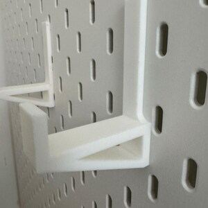 Op de afbeelding: Witte pegboard met twee witte plankbeugels. Het pegboard heeft gelijkmatig verdeelde, verticale ovale gaten. De L-vormige plankbeugels zijn ontworpen om in de gaten van het pegboard te passen en als steun te dienen.