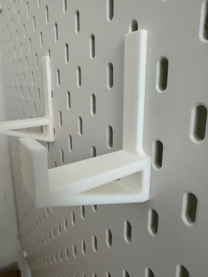Op de afbeelding: Witte pegboard met twee witte plankbeugels. Het pegboard heeft gelijkmatig verdeelde, verticale ovale gaten. De L-vormige plankbeugels zijn ontworpen om in de gaten van het pegboard te passen en als steun te dienen.