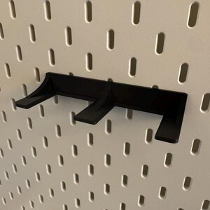 May include: A black pegboard shelf bracket is mounted on a white pegboard. The bracket has three angled supports for holding items. The pegboard has evenly spaced oval holes.