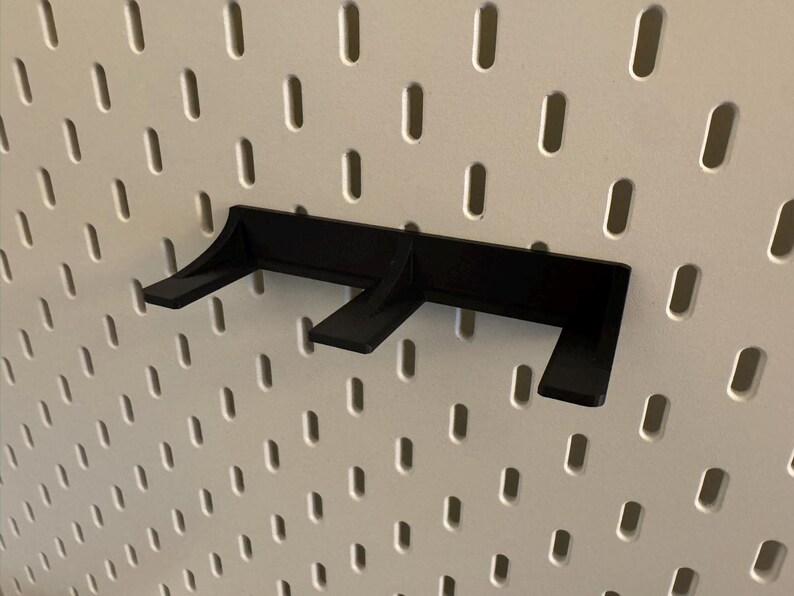 May include: A black pegboard shelf bracket is mounted on a white pegboard. The bracket has three angled supports for holding items. The pegboard has evenly spaced oval holes.