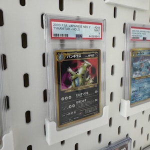 Peut inclure: Une carte Pok&eacute;mon class&eacute;e avec une carte Tyranitar-Holo de l'ensemble japonais Neo 2 de 2000. La carte est enferm&eacute;e dans un support de protection transparent et expos&eacute;e sur un panneau perfor&eacute; blanc. Le texte de la carte comprend "TYRANITAR-HOLO" et "MINT 9".
