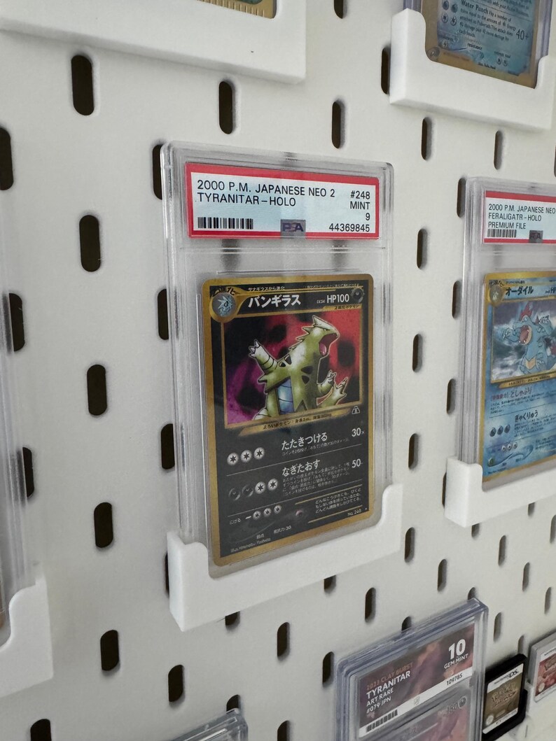 Peut inclure: Une carte Pok&eacute;mon class&eacute;e avec une carte Tyranitar-Holo de l'ensemble japonais Neo 2 de 2000. La carte est enferm&eacute;e dans un support de protection transparent et expos&eacute;e sur un panneau perfor&eacute; blanc. Le texte de la carte comprend "TYRANITAR-HOLO" et "MINT 9".