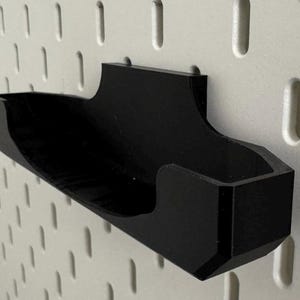 Op de afbeelding: Een zwarte plastic pegboardhaak met een gebogen ontwerp. De haak is gemonteerd op een wit pegboard.