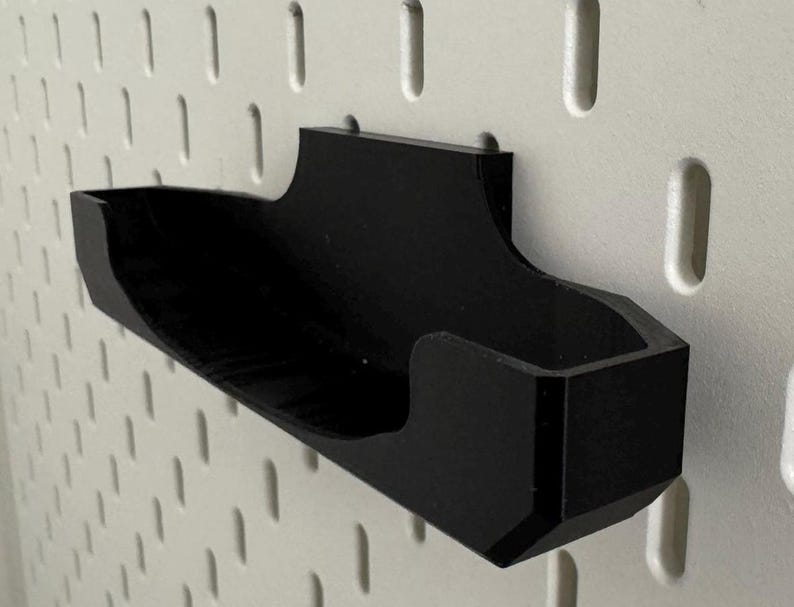 Op de afbeelding: Een zwarte plastic pegboardhaak met een gebogen ontwerp. De haak is gemonteerd op een wit pegboard.