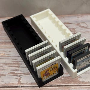 Puede incluir: Dos soportes para cartuchos de juegos impresos en 3D, uno negro y otro blanco, cada uno dise&ntilde;ado para contener m&uacute;ltiples cartuchos de juegos de Nintendo 3DS. Los soportes son rectangulares con ranuras individuales. Varios cartuchos de juegos son visibles.