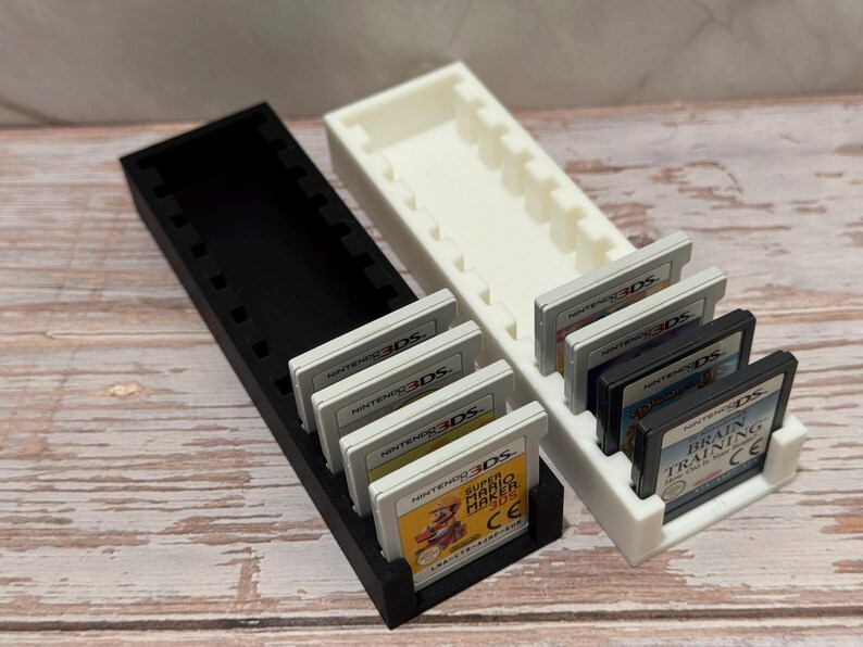 Puede incluir: Dos soportes para cartuchos de juegos impresos en 3D, uno negro y otro blanco, cada uno dise&ntilde;ado para contener m&uacute;ltiples cartuchos de juegos de Nintendo 3DS. Los soportes son rectangulares con ranuras individuales. Varios cartuchos de juegos son visibles.