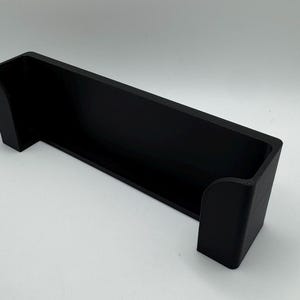 Pode incluir: Uma prateleira de parede de pl&aacute;stico preto com uma borda frontal curva. A prateleira &eacute; projetada para segurar um objeto pequeno, como um telefone ou um controle remoto.