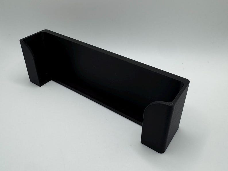 Pode incluir: Uma prateleira de parede de pl&aacute;stico preto com uma borda frontal curva. A prateleira &eacute; projetada para segurar um objeto pequeno, como um telefone ou um controle remoto.