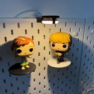 K&ouml;nnte beinhalten: Zwei Pop-Vinyl-Figuren, eine von einem M&auml;dchen mit roten Haaren und einem gr&uuml;nen Kleid und die andere von einem Jungen mit blonden Haaren und einer braunen Jacke, werden an einer wei&szlig;en Lochwand mit einem kleinen Licht dar&uuml;ber ausgestellt.