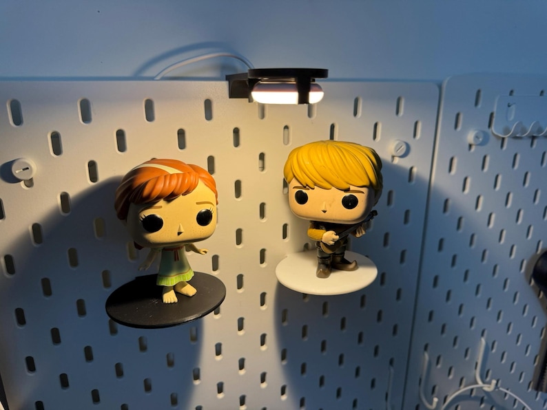 K&ouml;nnte beinhalten: Zwei Pop-Vinyl-Figuren, eine von einem M&auml;dchen mit roten Haaren und einem gr&uuml;nen Kleid und die andere von einem Jungen mit blonden Haaren und einer braunen Jacke, werden an einer wei&szlig;en Lochwand mit einem kleinen Licht dar&uuml;ber ausgestellt.