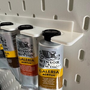 May include: A white pegboard organiser holds several tubes of Winsor & Newton Galeria acrylic paint. The paint tubes are silver with black caps and yellow and black labels. The organiser is made of white plastic.