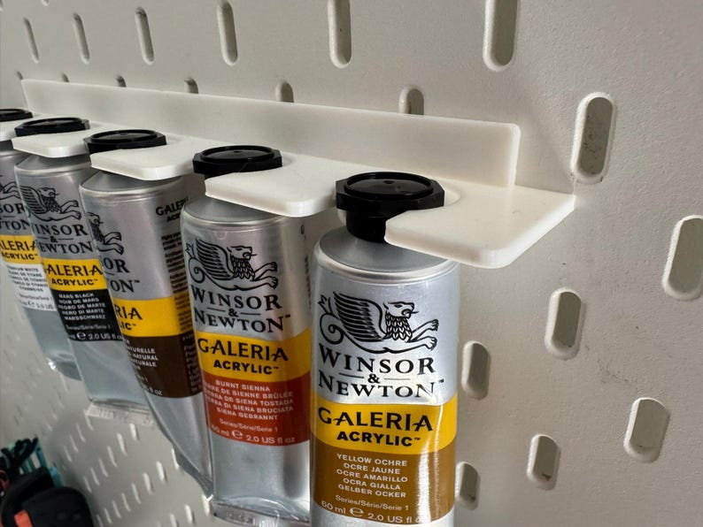May include: A white pegboard organiser holds several tubes of Winsor & Newton Galeria acrylic paint. The paint tubes are silver with black caps and yellow and black labels. The organiser is made of white plastic.