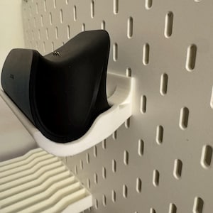 May include: A black electronic device rests on a white, curved shelf attached to a white pegboard. The shelf is designed to hold the device securely. The pegboard has evenly spaced oval holes.