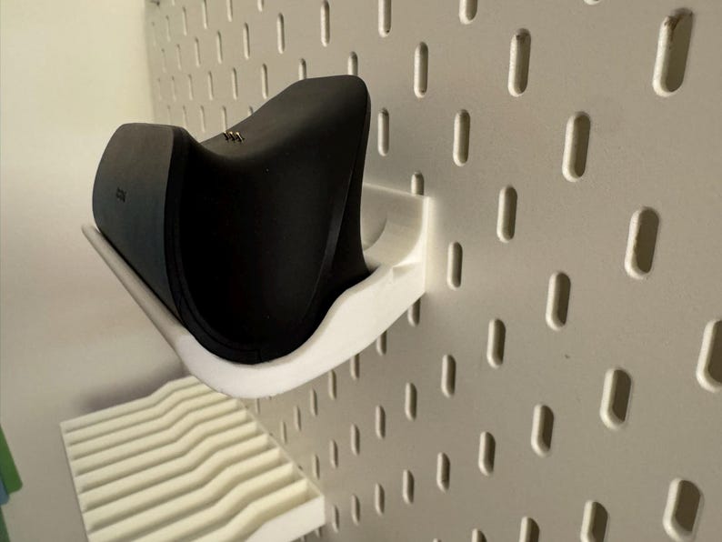 May include: A black electronic device rests on a white, curved shelf attached to a white pegboard. The shelf is designed to hold the device securely. The pegboard has evenly spaced oval holes.