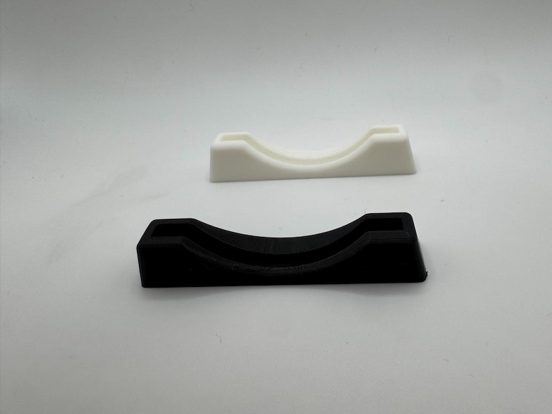 Puede incluir: Se muestran dos peque&ntilde;os soportes curvos, uno negro y otro blanco. Los soportes tienen un interior redondeado y una base rectangular. Parecen estar hechos de un material s&oacute;lido. Sobre fondo blanco.