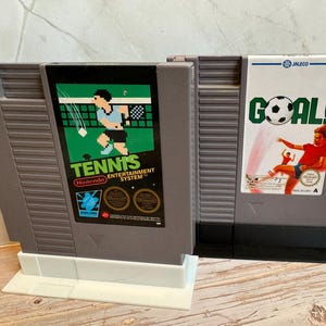 K&ouml;nnte beinhalten: Zwei Vintage-Spielmodule f&uuml;r das Nintendo Entertainment System. Eines zeigt eine Grafik eines Tennisspielers mit dem Text "Tennis Entertainment System". Das andere hat eine Grafik eines Fu&szlig;ballspielers mit dem Wort "GOAL!". Beide sind aus grauem Kunststoff.