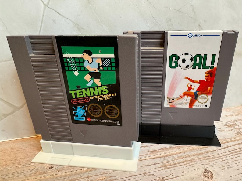 K&ouml;nnte beinhalten: Zwei Vintage-Spielmodule f&uuml;r das Nintendo Entertainment System. Eines zeigt eine Grafik eines Tennisspielers mit dem Text "Tennis Entertainment System". Das andere hat eine Grafik eines Fu&szlig;ballspielers mit dem Wort "GOAL!". Beide sind aus grauem Kunststoff.