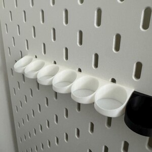 以下が含まれることがあります： 白のペグボードに、白と黒のカップ型のホルダーがいくつか取り付けられています。ペグボードには、等間隔に配置された細長い穴があります。ホルダーは、ペグボードの穴に収まるように設計されており、収納できます。