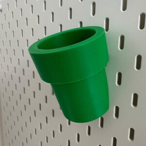 May include: A bright green, 3D-printed pipe, reminiscent of a video game, is angled against a white pegboard. The pipe has a cylindrical shape with a flared top and a curved bottom, suggesting a playful, decorative item.