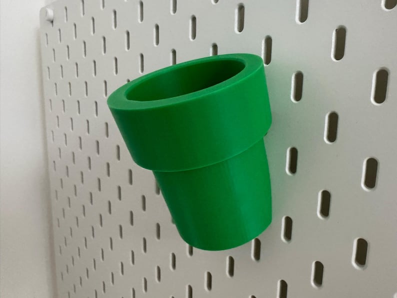May include: A bright green, 3D-printed pipe, reminiscent of a video game, is angled against a white pegboard. The pipe has a cylindrical shape with a flared top and a curved bottom, suggesting a playful, decorative item.