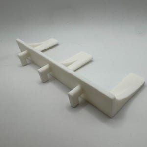 May include: A white plastic object with a rectangular base and several vertical supports. The object has a clean, minimalist design and appears to be a structural component or support for another item. The overall shape is angular and functional.