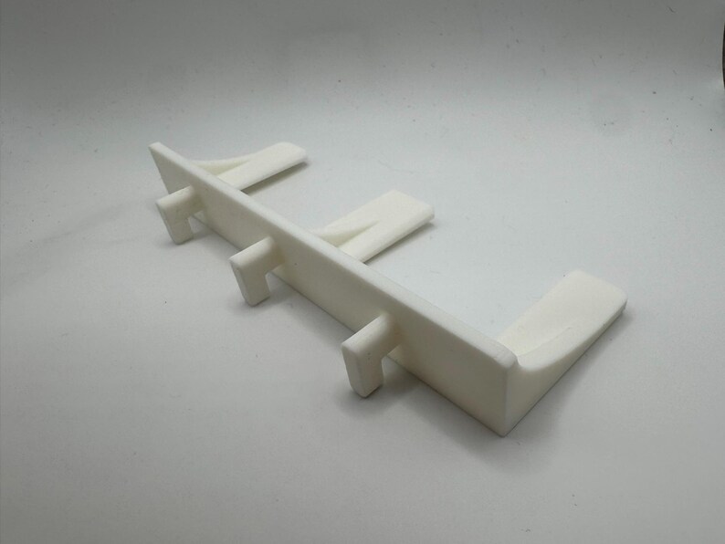 May include: A white plastic object with a rectangular base and several vertical supports. The object has a clean, minimalist design and appears to be a structural component or support for another item. The overall shape is angular and functional.