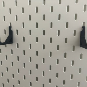 以下が含まれることがあります： 白いペグボードの壁に取り付けられた黒い金属製のペグボードフック2個
