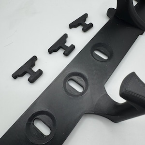 May include: Black 3D-printed plastic item with a long, flat base featuring three oval openings. Three smaller, T-shaped pieces are positioned nearby. The design includes curved elements, suggesting a mounting or support structure.