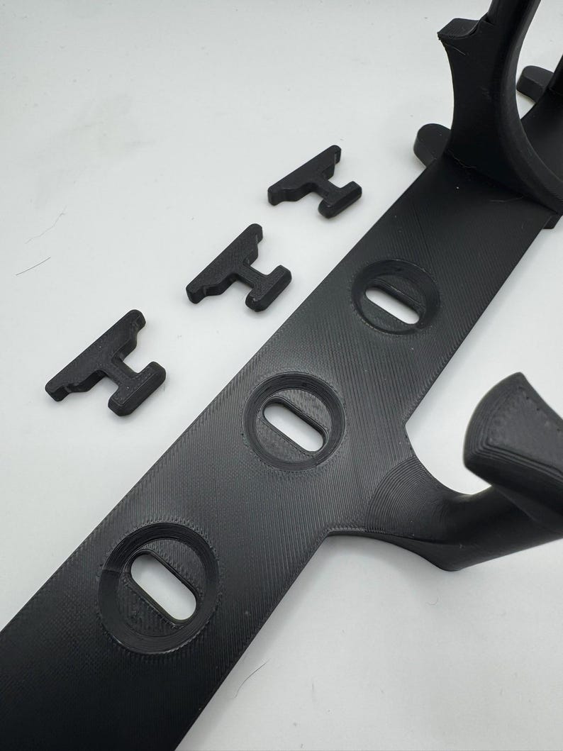 May include: Black 3D-printed plastic item with a long, flat base featuring three oval openings. Three smaller, T-shaped pieces are positioned nearby. The design includes curved elements, suggesting a mounting or support structure.