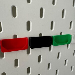 May include: Three small plastic storage bins, mounted on a white pegboard wall. The bins are red, black, and green.
