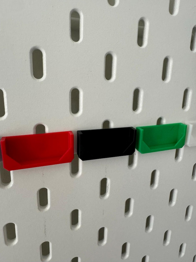 May include: Three small plastic storage bins, mounted on a white pegboard wall. The bins are red, black, and green.