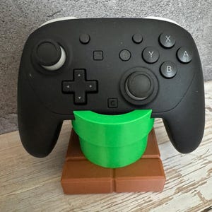 May include: A black video game controller rests on a green pipe-shaped stand, which is set on a brown brick-like base. The controller has white accents and various buttons. The stand is designed to hold the controller.