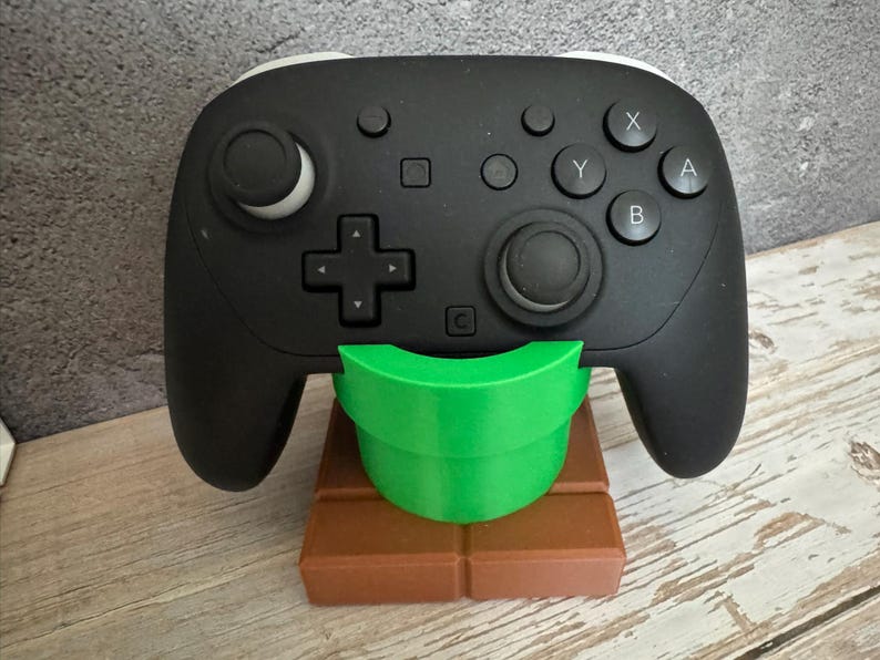 K&ouml;nnte beinhalten: Ein schwarzer Videospiel-Controller liegt auf einem gr&uuml;nen, rohrf&ouml;rmigen St&auml;nder, der auf einer braunen, ziegelartigen Basis steht. Der Controller hat wei&szlig;e Akzente und verschiedene Tasten. Der St&auml;nder ist zur Aufnahme des Controllers konzipiert.