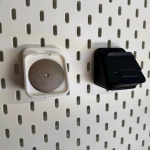 May include: Two wall-mounted storage accessories. One is white with a silver disc, the other is black. Both are attached to a white pegboard with oval-shaped holes. The accessories appear to be designed for holding small items.