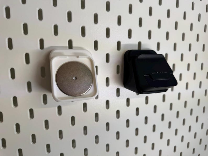 May include: Two wall-mounted storage accessories. One is white with a silver disc, the other is black. Both are attached to a white pegboard with oval-shaped holes. The accessories appear to be designed for holding small items.