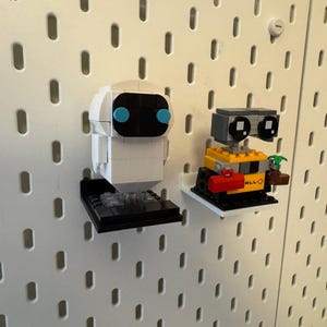 May include: Two white and yellow Lego robot figures, one with blue eyes and the other with black eyes, are displayed on a white pegboard wall. The robots are on black and white bases.