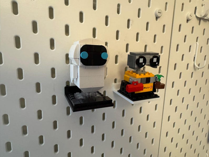 May include: Two white and yellow Lego robot figures, one with blue eyes and the other with black eyes, are displayed on a white pegboard wall. The robots are on black and white bases.