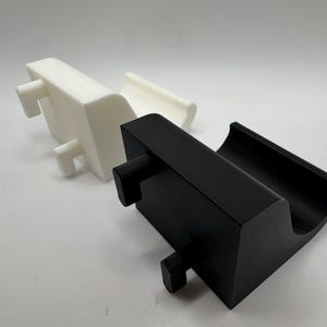 K&ouml;nnte beinhalten: Zwei 3D-gedruckte Telefonst&auml;nder. Einer ist wei&szlig;, der andere ist schwarz. Jeder St&auml;nder hat eine rechteckige Basis mit einer gebogenen Oberseite, um ein Telefon zu halten, und zwei hervorstehende Stifte f&uuml;r Stabilit&auml;t.