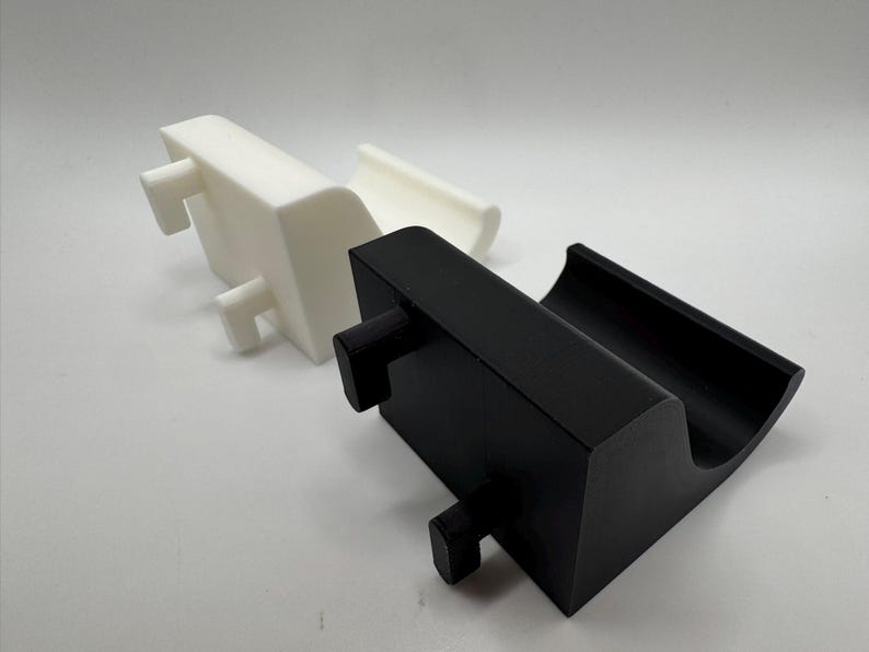 K&ouml;nnte beinhalten: Zwei 3D-gedruckte Telefonst&auml;nder. Einer ist wei&szlig;, der andere ist schwarz. Jeder St&auml;nder hat eine rechteckige Basis mit einer gebogenen Oberseite, um ein Telefon zu halten, und zwei hervorstehende Stifte f&uuml;r Stabilit&auml;t.