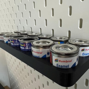 May include: A black shelf holds several small paint cans with silver lids. The cans are labeled "Humbrol Enamel" and "Revell Email Color." The shelf is mounted on a white pegboard.