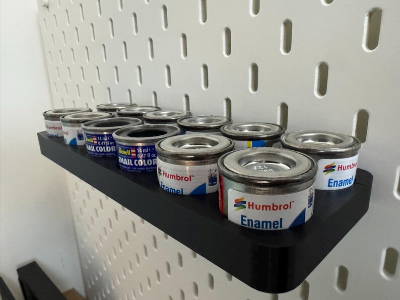 May include: A black shelf holds several small paint cans with silver lids. The cans are labeled "Humbrol Enamel" and "Revell Email Color." The shelf is mounted on a white pegboard.