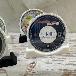 Puede incluir: Soportes de pl&aacute;stico blanco para discos UMD que muestran varios discos de juegos. El disco m&aacute;s frontal est&aacute; etiquetado como "Wipeout Pure" con el texto "UMD Universal Media Disc". Otros discos incluyen t&iacute;tulos de "Grand Theft Auto: Liberty City Stories" y "Star Wars".