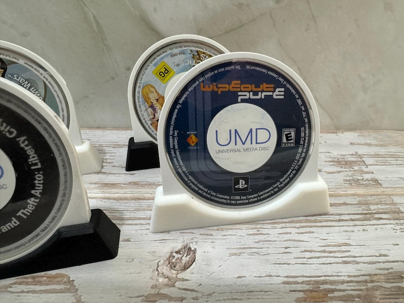 Puede incluir: Soportes de pl&aacute;stico blanco para discos UMD que muestran varios discos de juegos. El disco m&aacute;s frontal est&aacute; etiquetado como "Wipeout Pure" con el texto "UMD Universal Media Disc". Otros discos incluyen t&iacute;tulos de "Grand Theft Auto: Liberty City Stories" y "Star Wars".