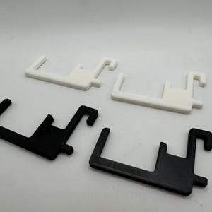 May include: Four plastic, hook-shaped objects. Two are white, and two are black. The objects have a rectangular base with a hook extending from one end and a small peg on the side. The items are likely 3D printed.