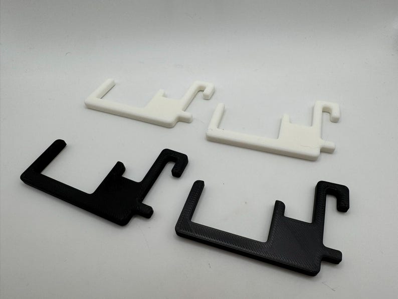May include: Four plastic, hook-shaped objects. Two are white, and two are black. The objects have a rectangular base with a hook extending from one end and a small peg on the side. The items are likely 3D printed.